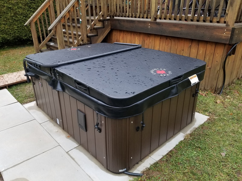 Hard-shell hot tub with a rain-speckled Canadian Spa Co. cover and brown cabinet on a paved base beside a timber deck