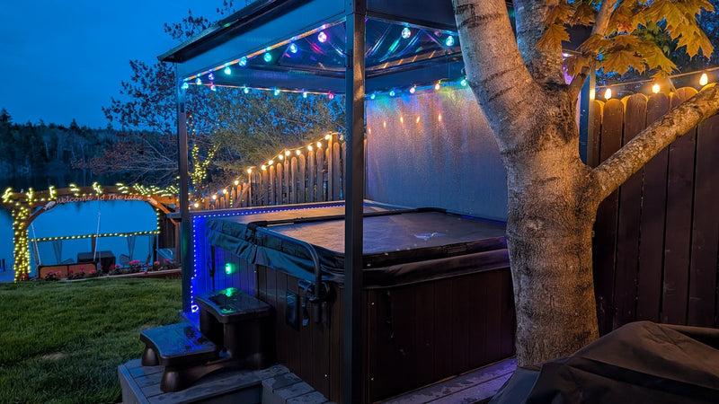 Hard-shell hot tub with its cover closed beside a garden shelter with string lights, spa steps and an autumn tree at dusk