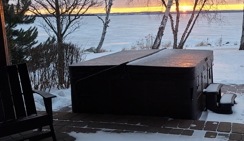 Hot tub with its cover closed and steps alongside on a snowy patio at dusk, with birch trees and a frozen lake behind