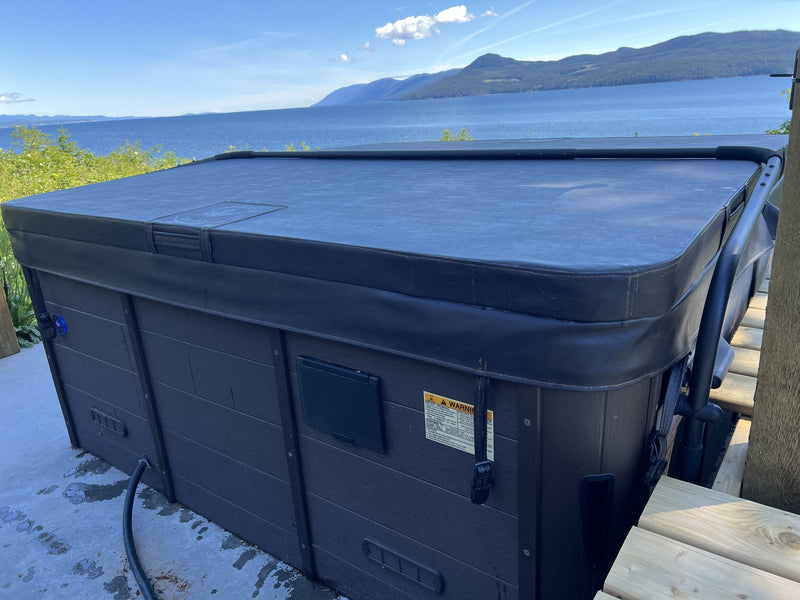 Hard-shell hot tub with the cover closed and strapped down beside a timber deck, with a lake and hills behind