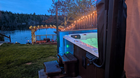 
Hot tub lit blue and green at dusk with the cover folded back on a lifter, steps beside it and string lights along the garden