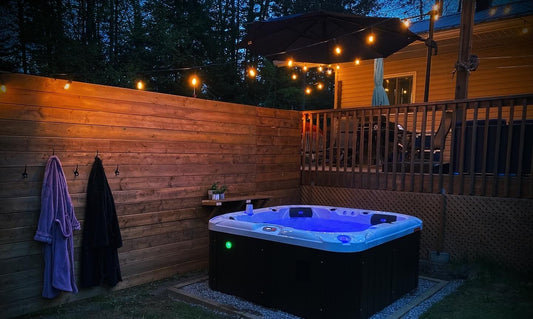 
Hard-shell hot tub lit blue at dusk beside a timber fence, with two bathrobes hanging on hooks