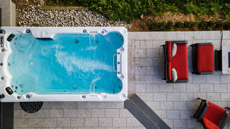 Aerial view of a St Lawrence 13ft swim spa with its jets running, beside a tiled patio with red-cushioned garden seating