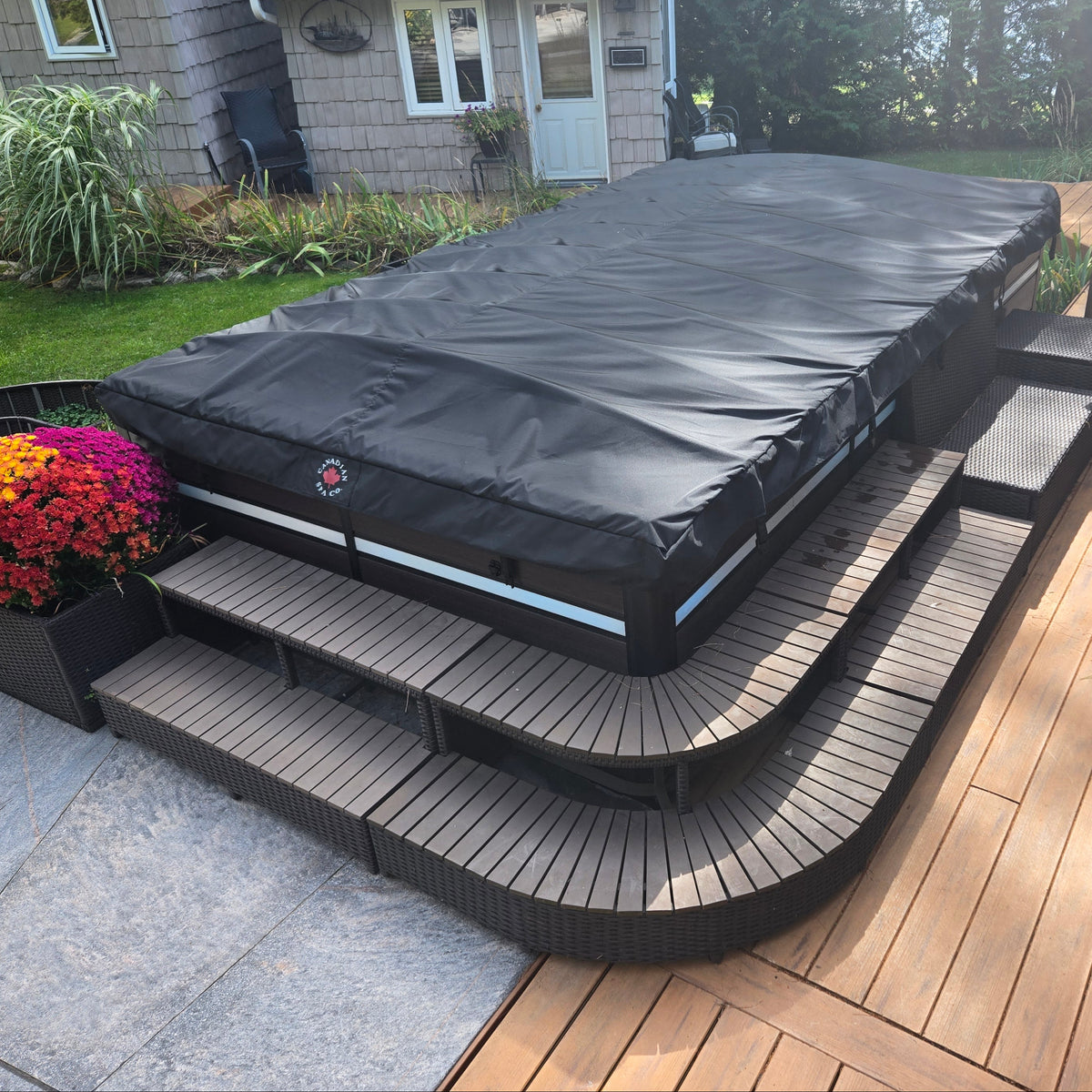 
A black hot tub cover with a small red and white logo patch sits on a three-step wooden platform beside a wood deck, with a h
