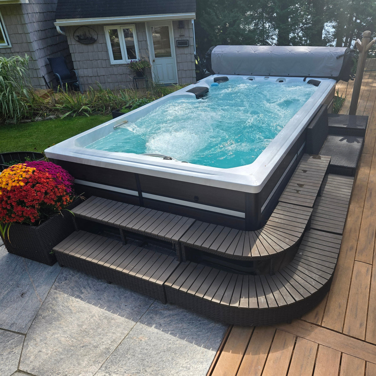 
An outdoor hot tub with bubbling turquoise water, set on a curved dark wood platform next to a wooden deck and house yard.