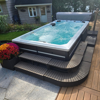 An outdoor hot tub with bubbling turquoise water, set on a curved dark wood platform next to a wooden deck and house yard.