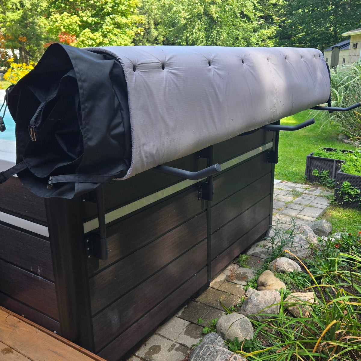
A dark brown wooden hot tub with a light gray tufted cover partially rolled up, set in a backyard garden next to a stone walk