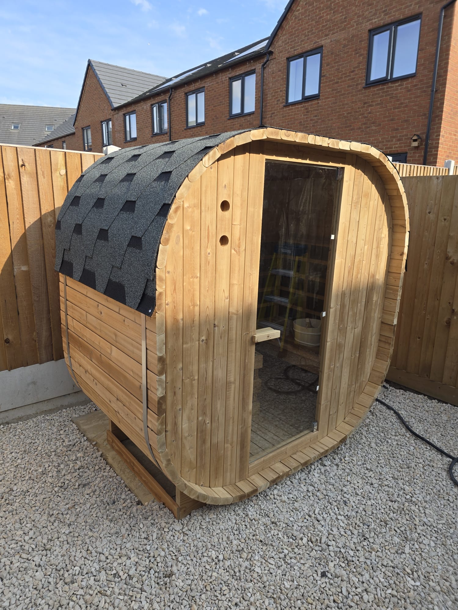 Outdoor Oval Barrel sauna set onto existing flagstones with a brick house and gravel base behind