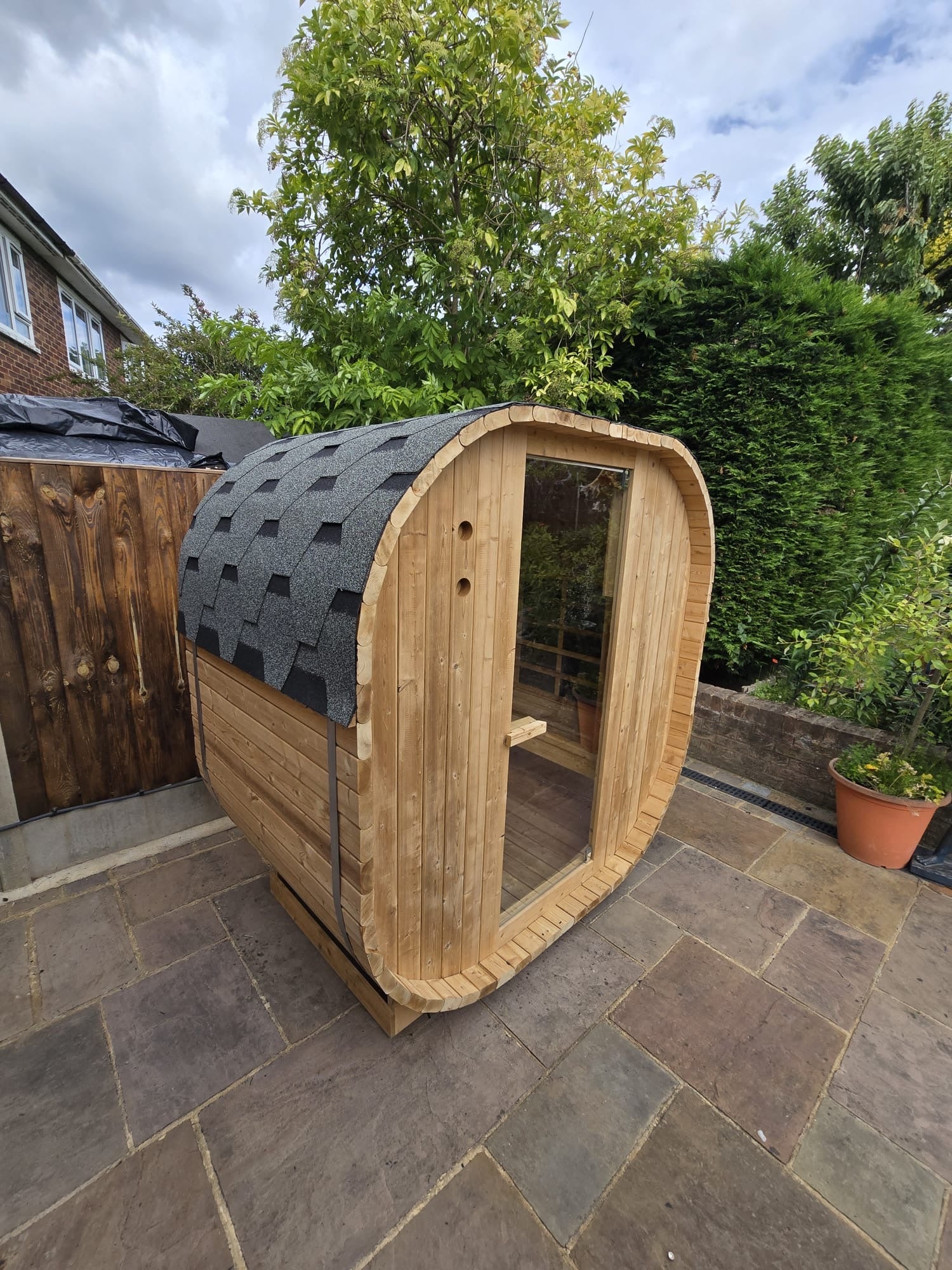 Outdoor Oval Barrel sauna with a full-glass door, settled onto an existing brick path at the edge of a woodland boundary