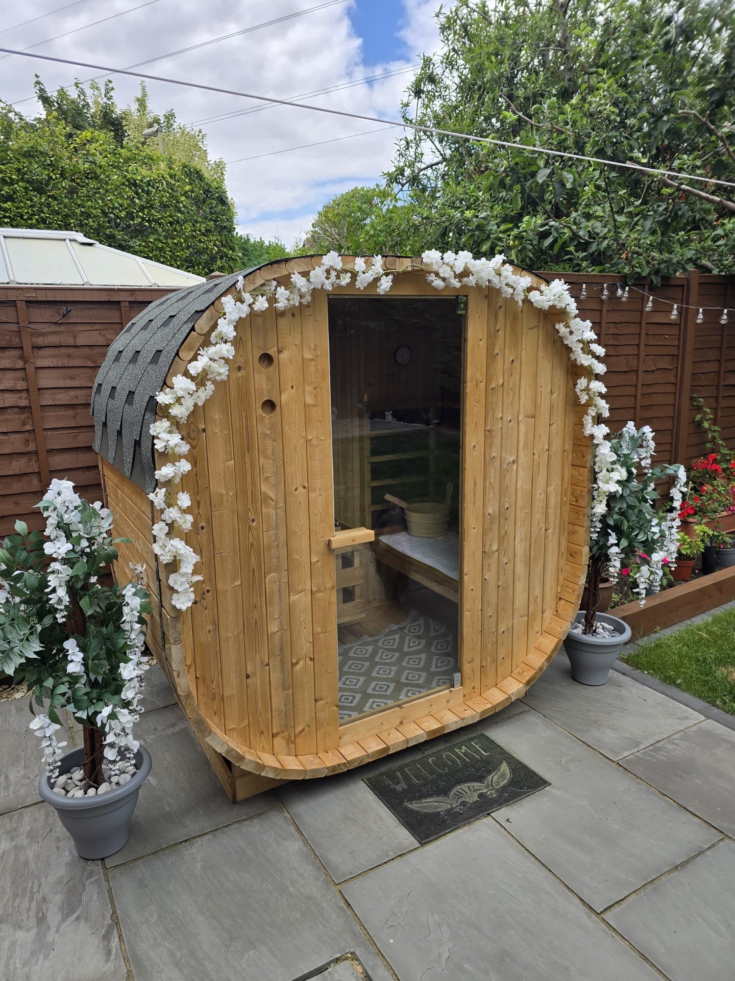 Outdoor Oval Barrel sauna on a slate patio in an urban garden, open-glass front looking through to the lawn beyond