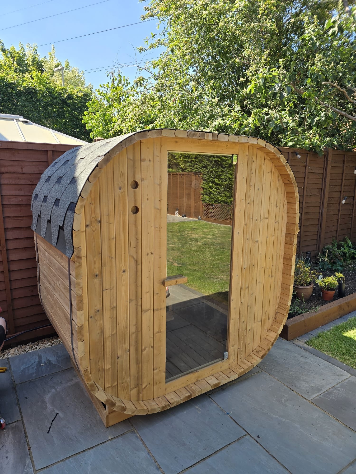 Outdoor Oval Barrel sauna on a compacted gravel pad against the boundary fence in a new-build garden