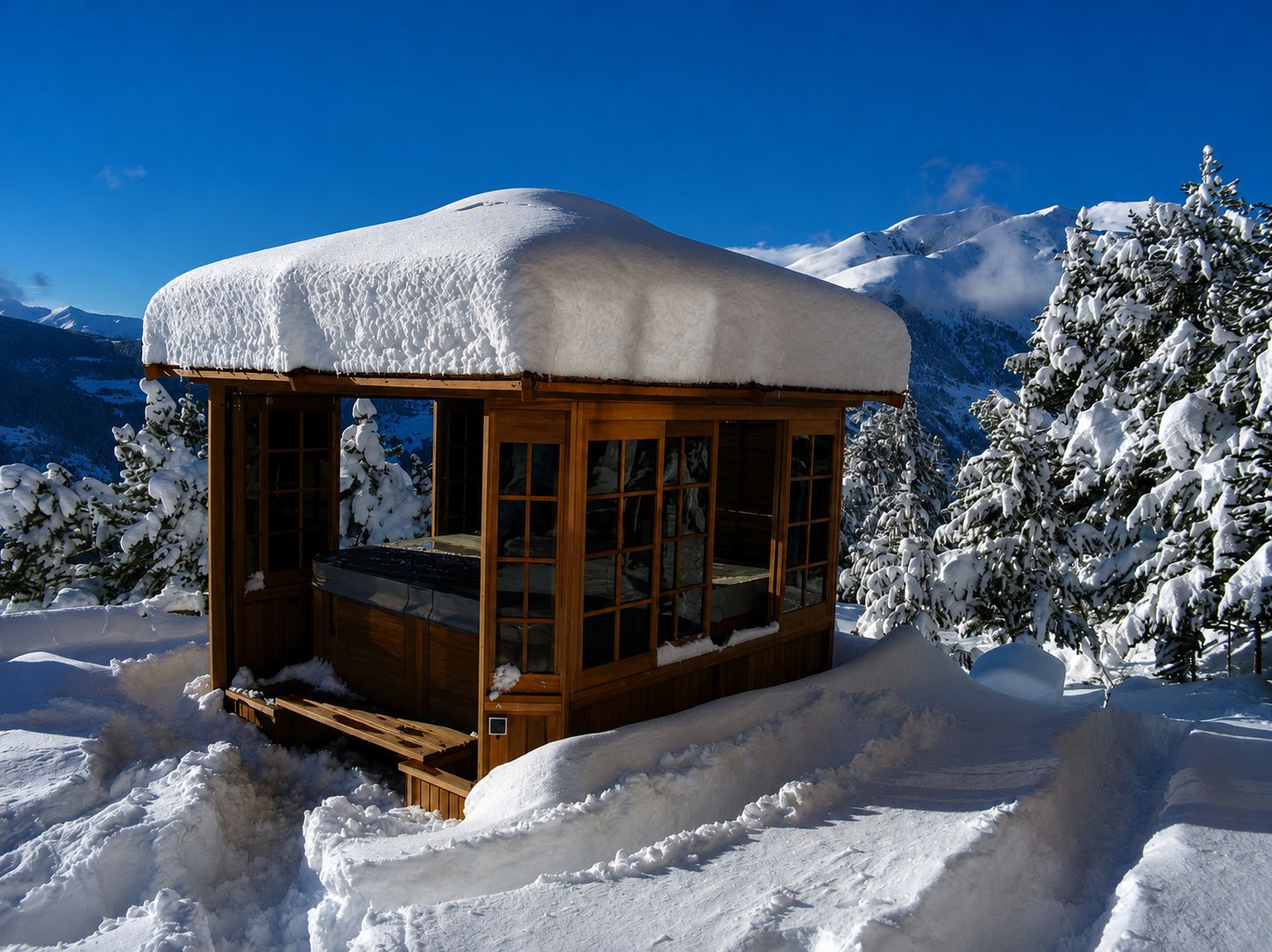 Snow-covered wooden gazebo sheltering a covered hot tub, with snow-laden pine trees and mountains under a clear blue sky